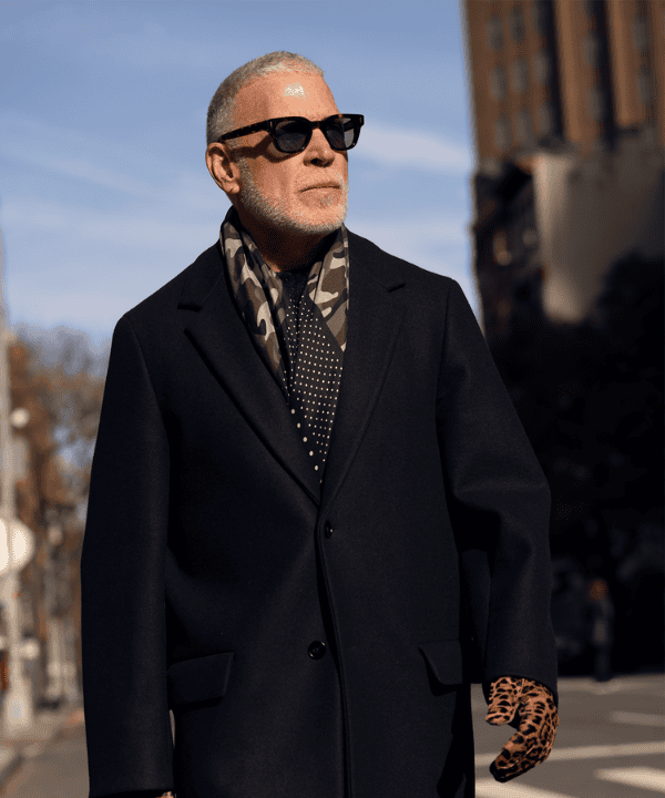 Free agent, Nick Wooster wearing long blakck trench coat and silk camoflauge scarf around neckwith sunglasses