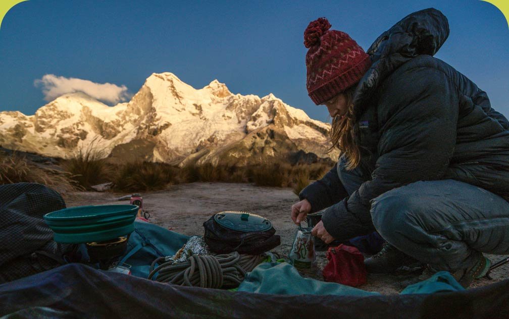 A climber cooks her breakfast on a camp stove with a large snowy mountain behind her catching first light of the day.