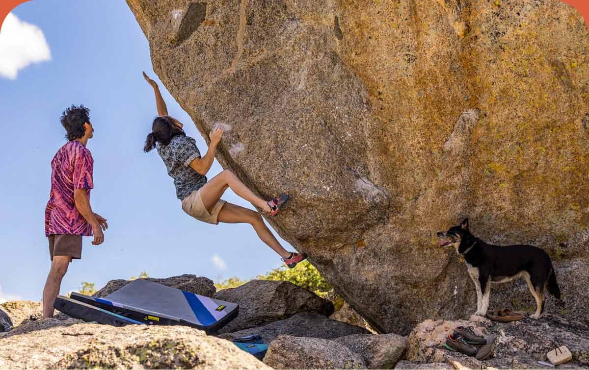 One person climbing on a big boulder while the other person watches. 