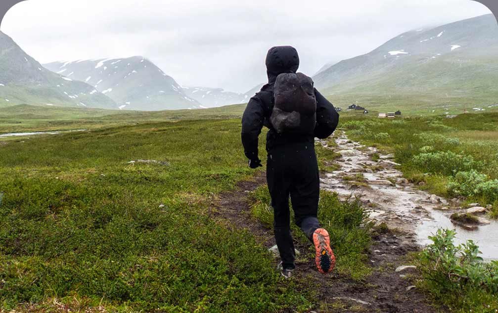 A person wearing rain gear and an backpack runs through a grassy valley bordered by mountains.