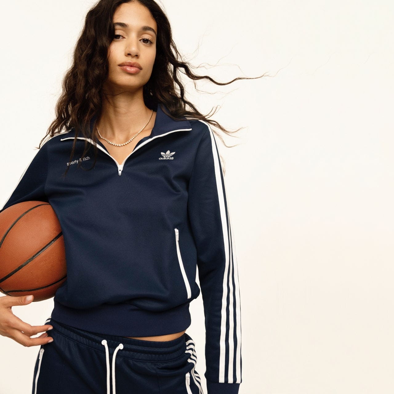 A model wearing a navy adidas Originals x Sporty & Rich track top and holding a basketball poses against a cream wall.