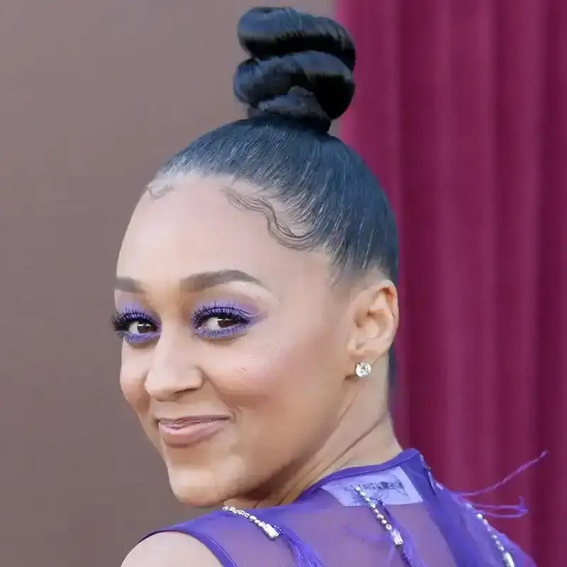 Tia Mowry appears on the red carpet in a purple dress in a high bun hairstyle and purple eye shadow.