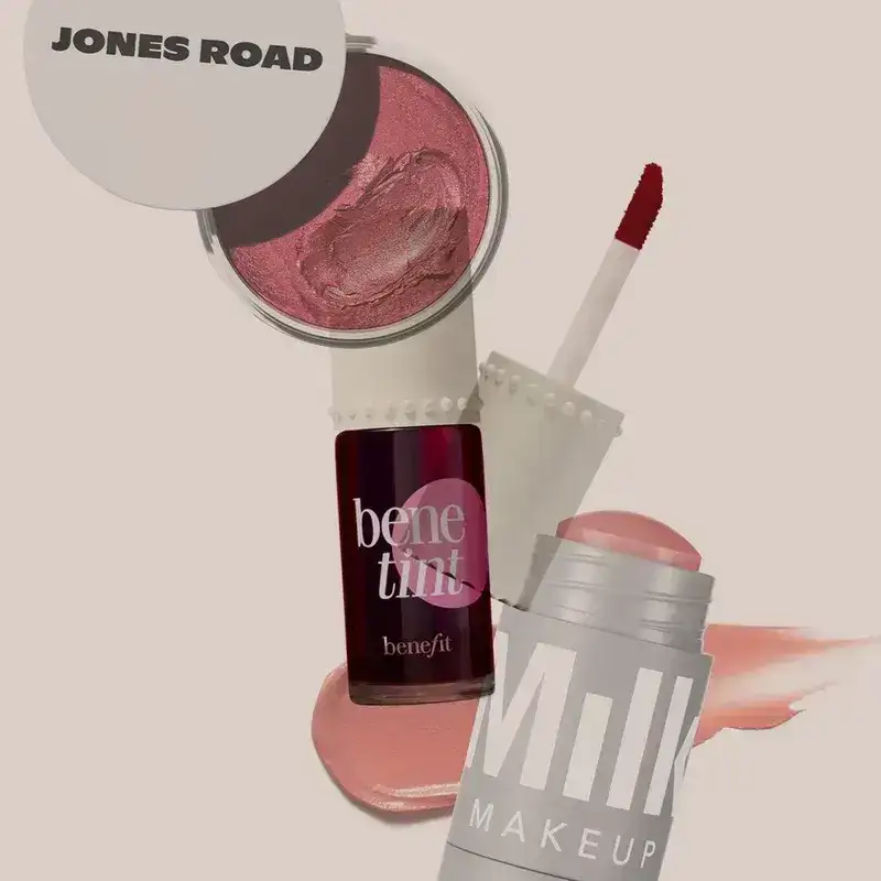 3 cosmetic items overlapping on a beige background. From top to bottom: small tin of cream, bottle with brush, and tube with stick.