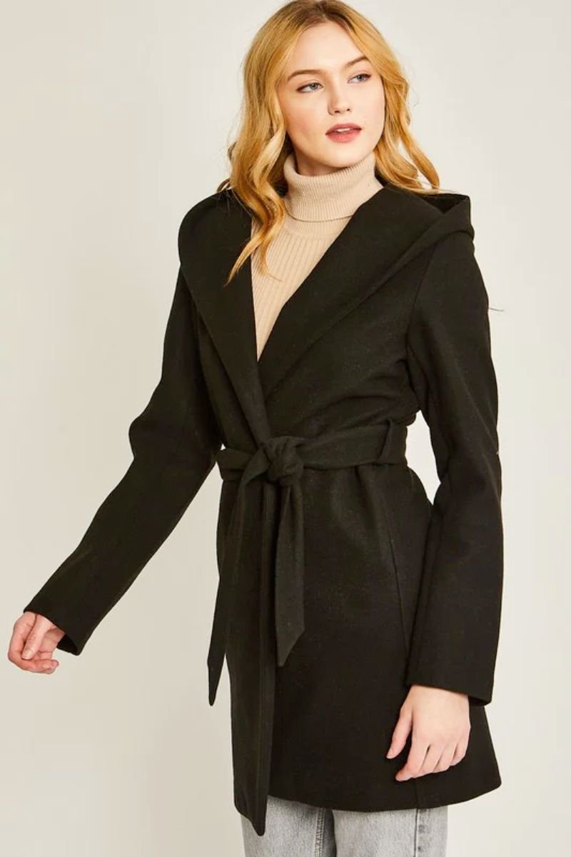 JQ Fleece Belted Hoodie Coat. The model is wearing a black coat and a tan turtleneck shirt underneath. The coat has a tie and the model has blonde hair. She is also wearing grey jeans.