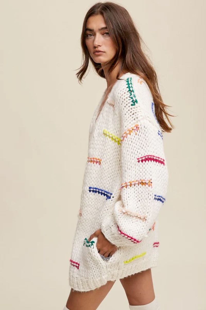 Hand Crochet Knit Stripe Design Open Cardigan. The model is wearing a cream colored cardigan with colorful stitched lines. She is also wearing tall white socks.