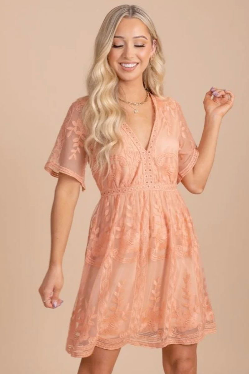 Light My Fire Peach Lace Mini Dress. The model is wearing a peach mini dress with see through sleeves. The model has gold jewelry and blonde hair.