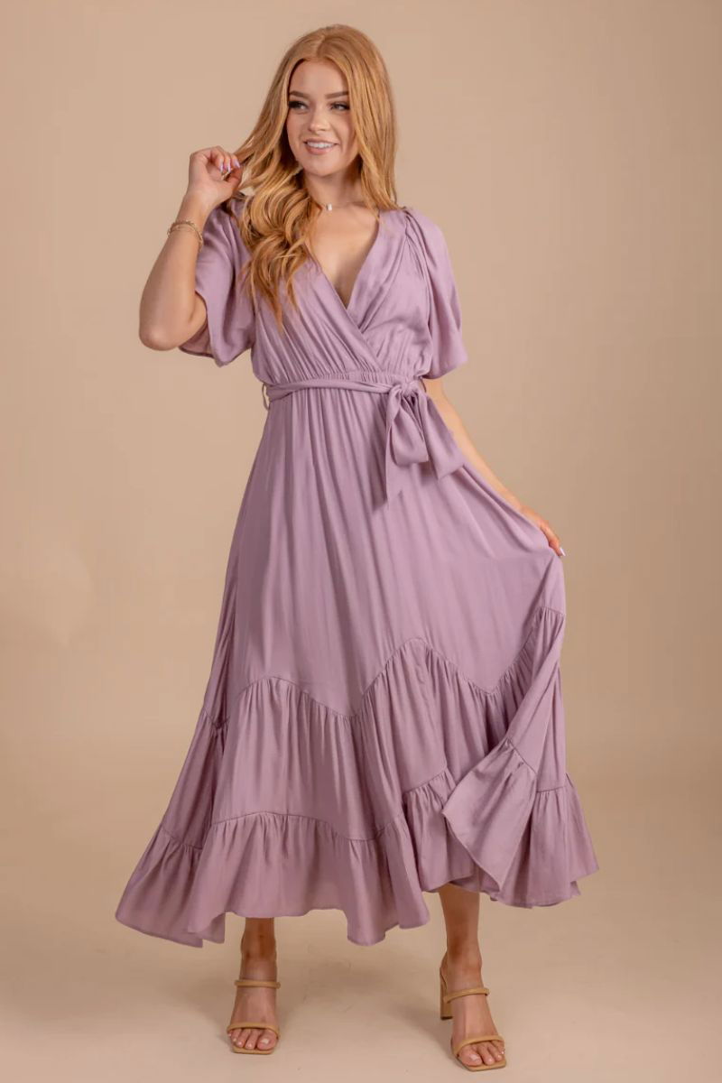 Sweetest Kiss Maxi Dress. The model is wearing a purple dress with a flowing silhouette and a tiered skirt. There is a waist tie and a v neckline. She is wearing tan heels.