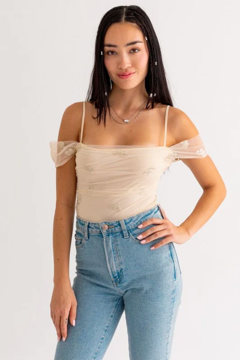 Embroidered Mesh Bodysuit. The woman is wearing a tan bodysuit with off the shoulder sleeves and blue jeans.