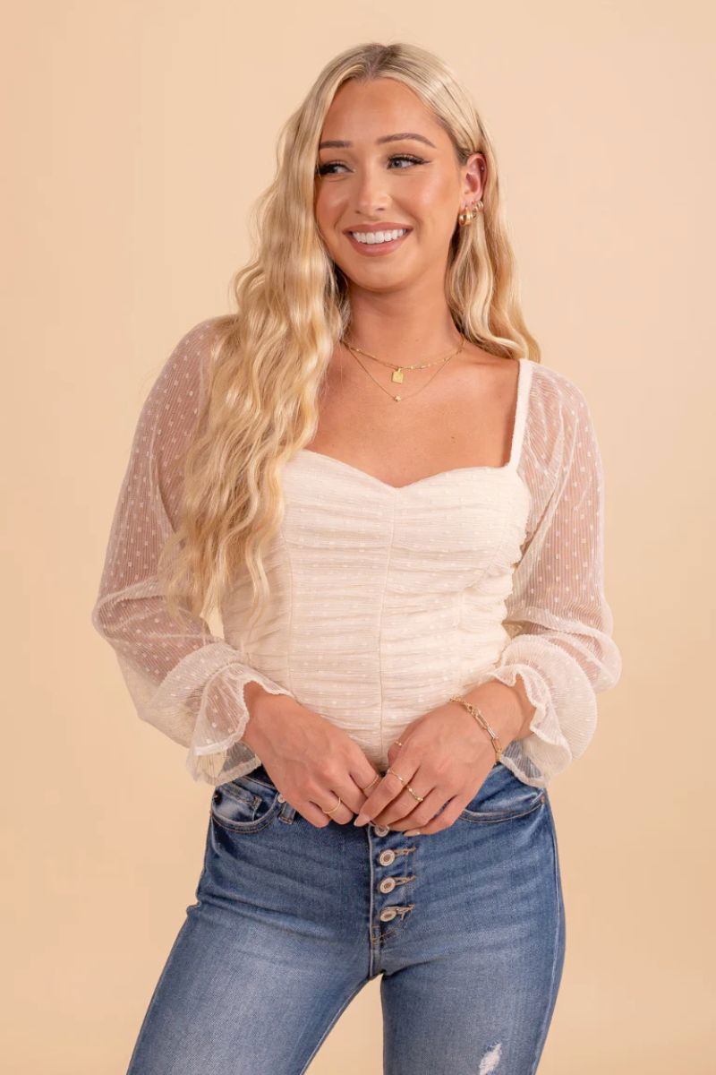 Sweet Romance Sheer Accent Long Sleeve Bodysuit. The model is wearing a ruched cream colored bodysuit with see through sleeves with dot details. The model is also wearing blue jeans.