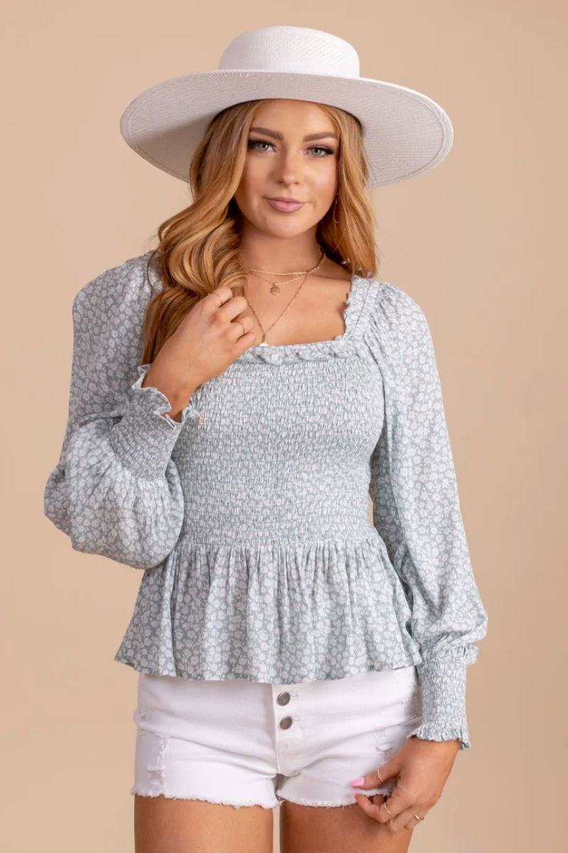 Desert Wildflower Smocked Long Sleeve Top. The model is wearing a light blue floral top with a ruched bodice and cuffed sleeves. She is also wearing a white wide brimmed hat and white shorts.