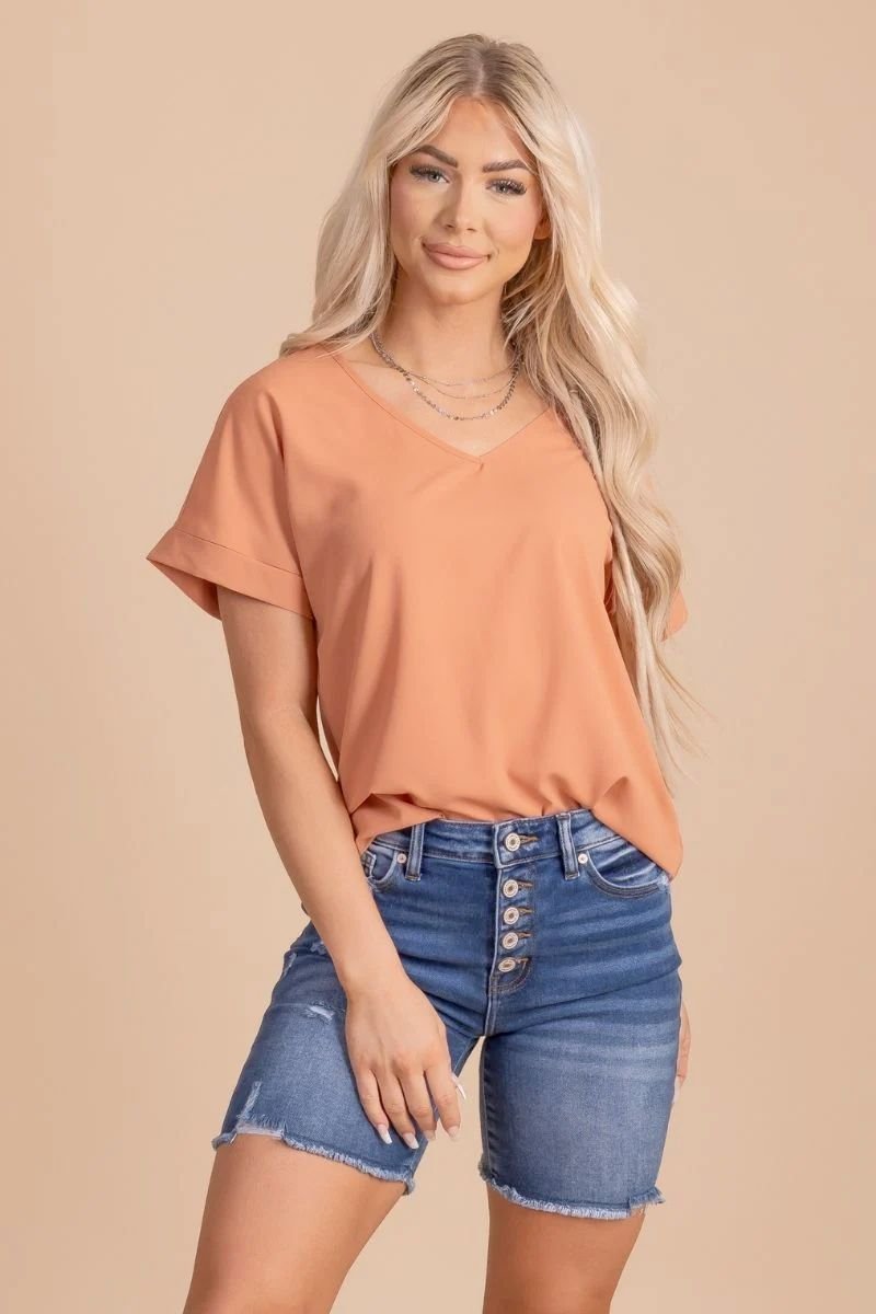 First Kiss V-Neck Blouse. The model is wearing a light orange shirt with jewelry and blue jeans.