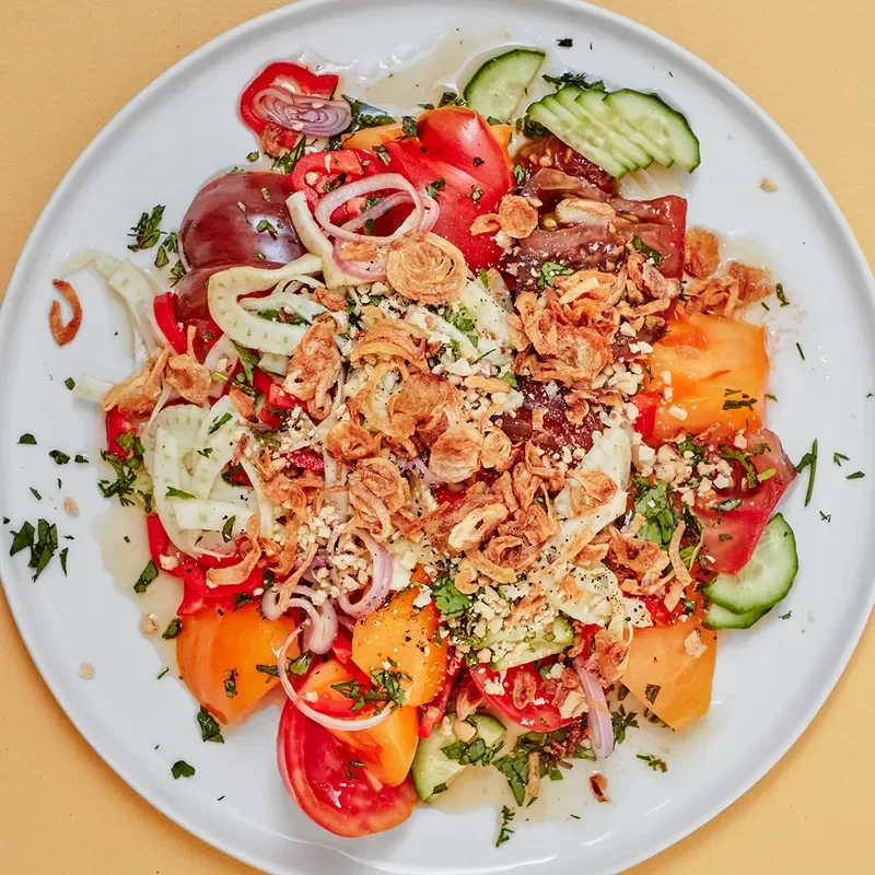 A white plate with multi-colored yellow, red, and purple tomato slices topped with crispy onions, cucumbers, and herbs, on a muted yellow background.