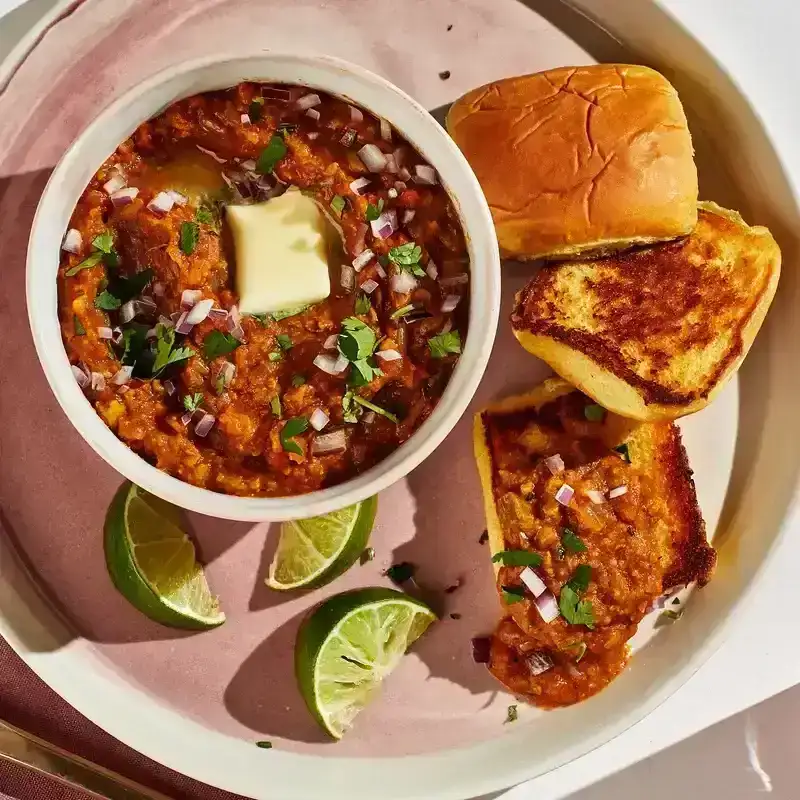 Bowl of stew sitting on a plate with buns and lime slices.