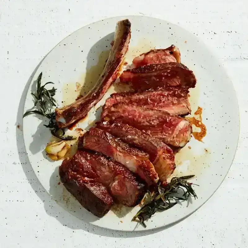 Rib eye steak on a plate with spent garlic and rosemary. The steak has been sliced away from the bone and cut into strips, revealing the blush interior.