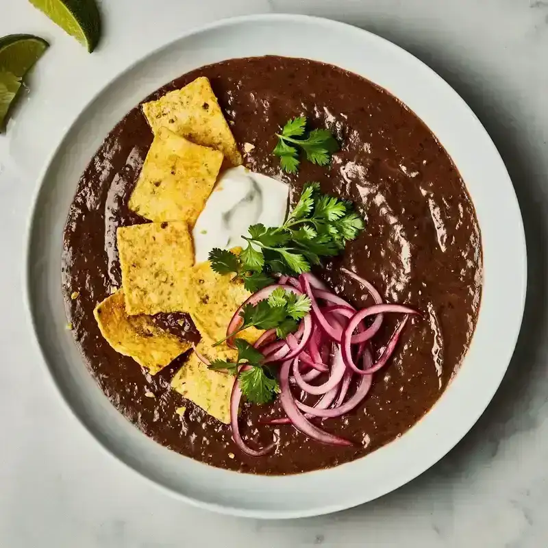 A bowl of silky black bean soup topped with sour cream, corn chips, cilantro, and pickled red onions.