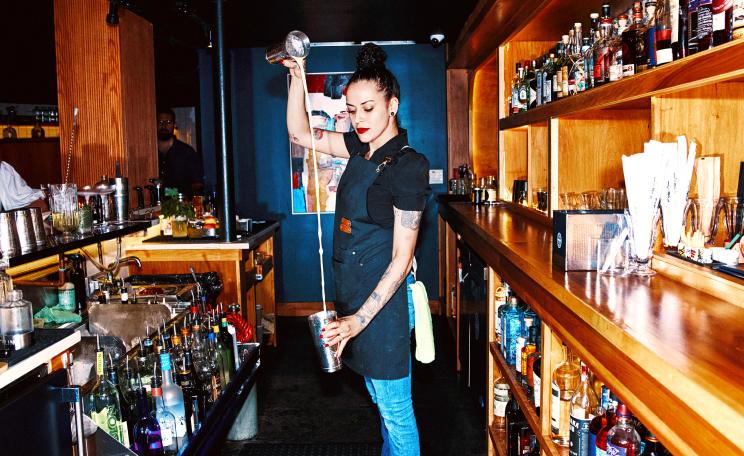 A bartender pours a drink from a shaker into a glass behind a bar stocked with various bottles and glasses.