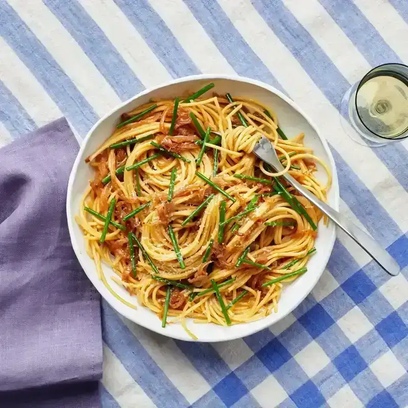 Bowl of Jammy Onion and Miso Pasta on blue gingham tablecloth with glass of white wine.