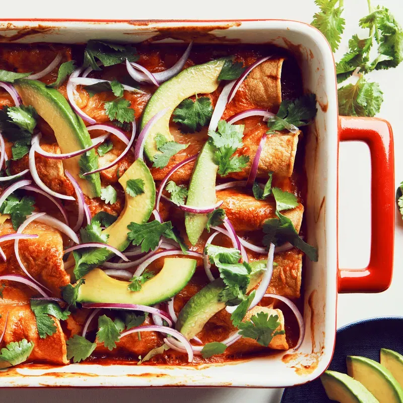 Beans, red and green bell peppers, and tortillas baked in a casserole dish and topped with avocados, red onions, and cilantro.