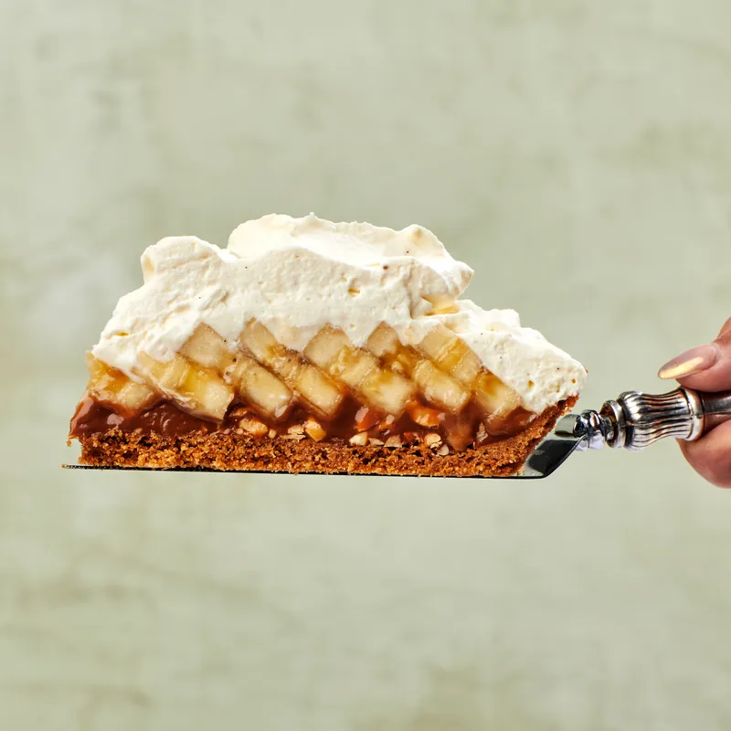 Side view of a slice of banoffee pie, with a peanut caramel, shingled banana slices, and a mound of cream, balanced on a pie server 