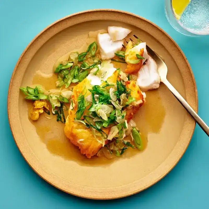 A crusty cod fillet on a plate, topped with a butter-scallion relish, with a pool of sauce gathering on the plate.