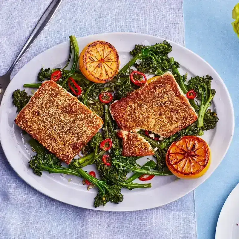 Two blocks of sesame feta sitting on a platter of broccolini with charred lemons and red peppers 