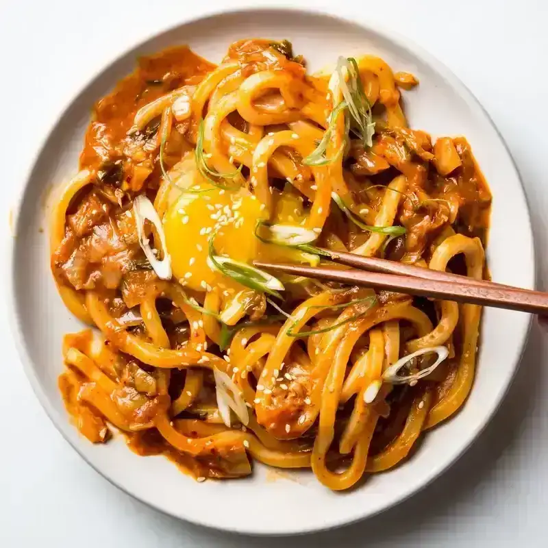 A white plate of kimchi udon noodles with a pair of chopsticks breaking a yellow egg yolk