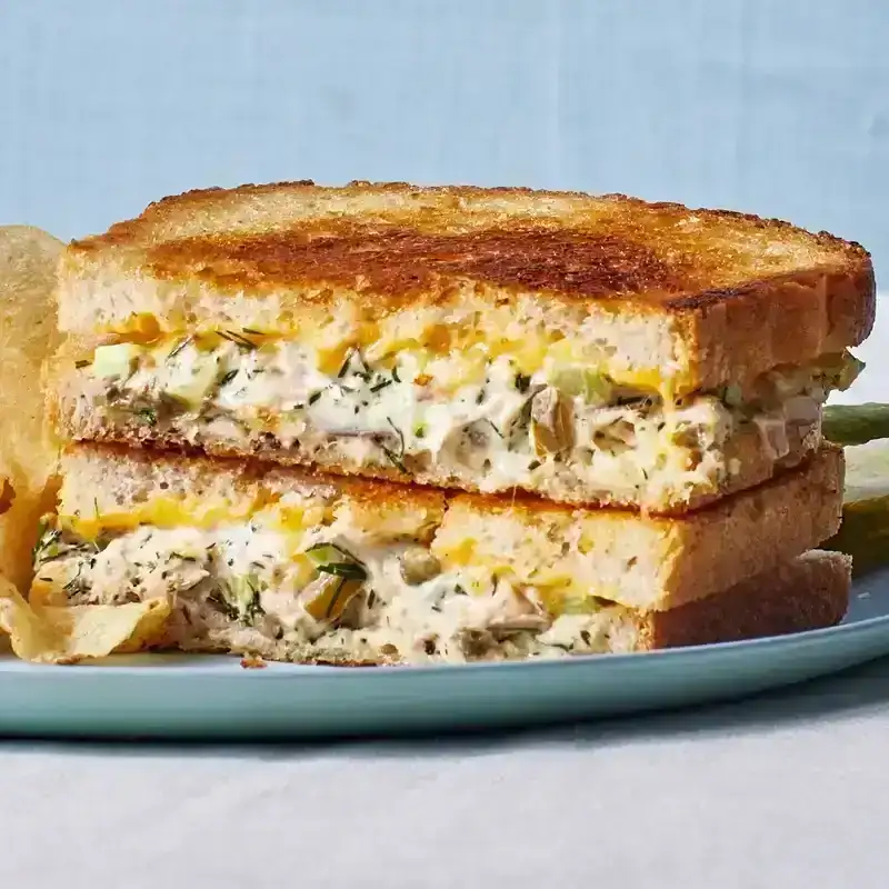 Two halves of a classic tuna melt sandwich stacked on a plate with potato chips covered in Frank's red hot and a pickle on the side.