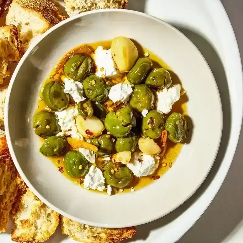 Marinated crumbles of feta cheese and Castelvetrano olives in a bowl with garlic and red pepper flakes alongside cubes of bread for dipping.