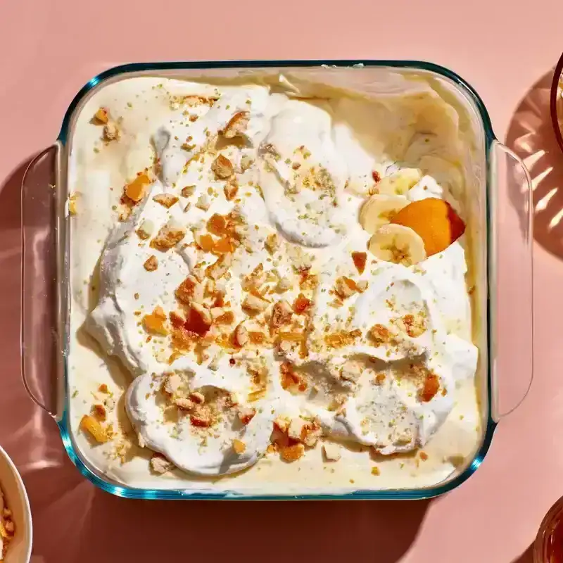 An 8x8 dish of banana pudding with a whipped cream topping and crushed vanilla wafers on top, alongside several serving bowls with scoops of pudding.