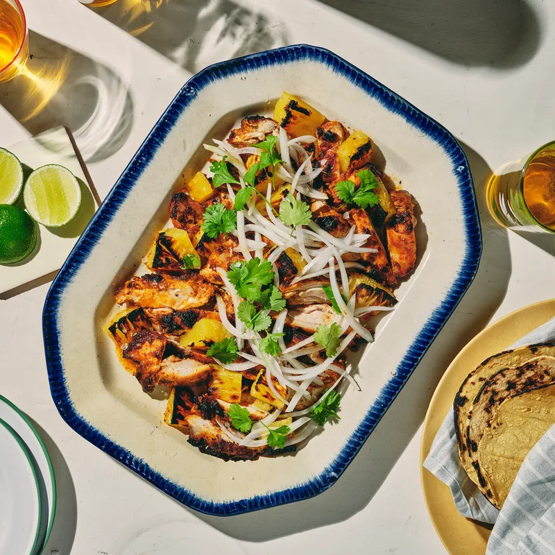 A platter of al pastor chicken garnished with cilantro and onions, next to a plate of tortillas.