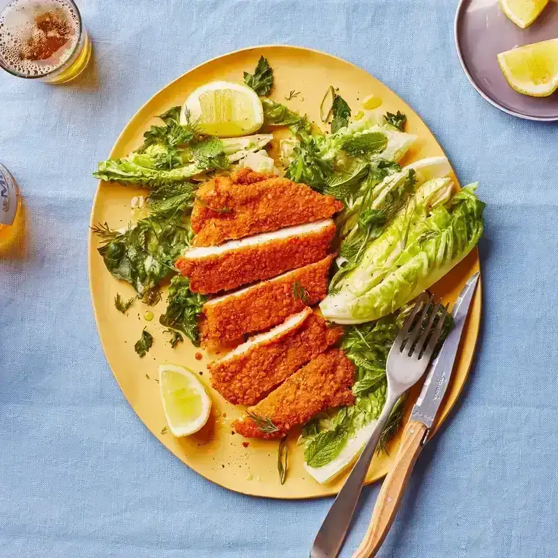 A plate of sliced crispy chicken cutlet with side salad on blue tablecloth. 