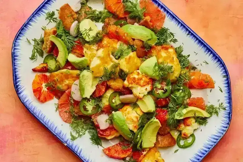 A citrus, Halloumi, and Avocado on a white and blue plate placed on a pink pastel surface;