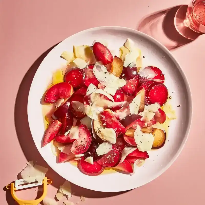 Large plated fruit salad with sliced plums and jagged Parmesan shavings, topped with olive oil and black pepper.