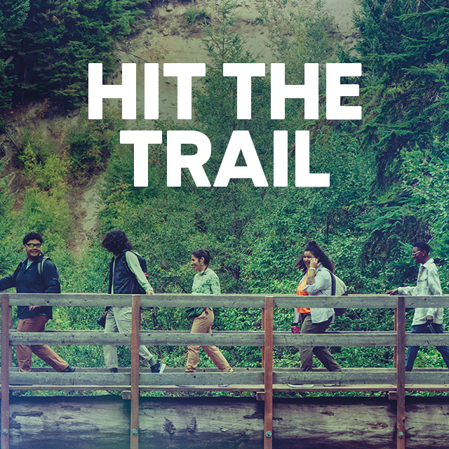 A group of people hiking cross a bridge with lush greenery behind them.