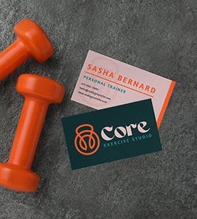 Two business cards for Core Exercise Studio employees rest on a concrete surface next to dumbbells.