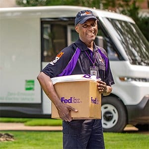 A smiling purple shirted FedEx team member holds a package.