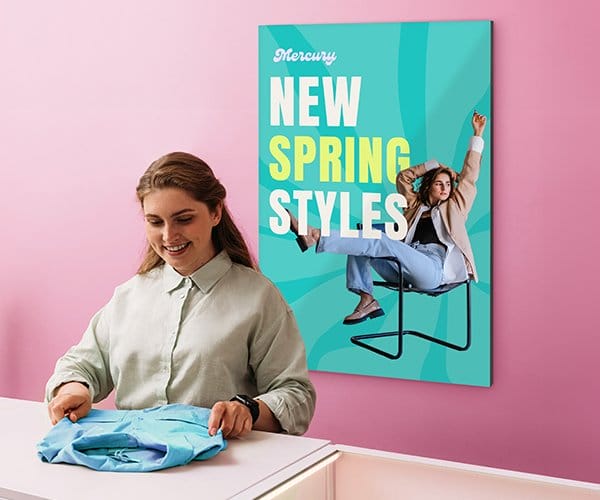 A woman folding a shirt at a clothing store and a photo poster hangs behind her that reads “NEW SPRING STYLES.”