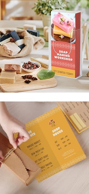 A stack of brochures in a plastic holder next to some handmade soap advertises a soap making course. Below, a folded yellow brochure advertising business hours lays open on a wooden surface.