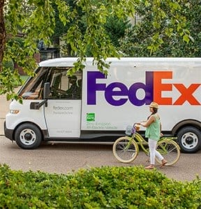 An electric FedEx Office vehicle drives down the road next to a person on a bicycle. 