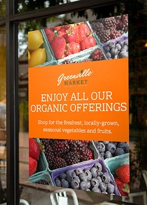 A colorful poster print displayed in a storefront window reads “ ENJOY ALL OUR ORGANIC OFFERINGS”