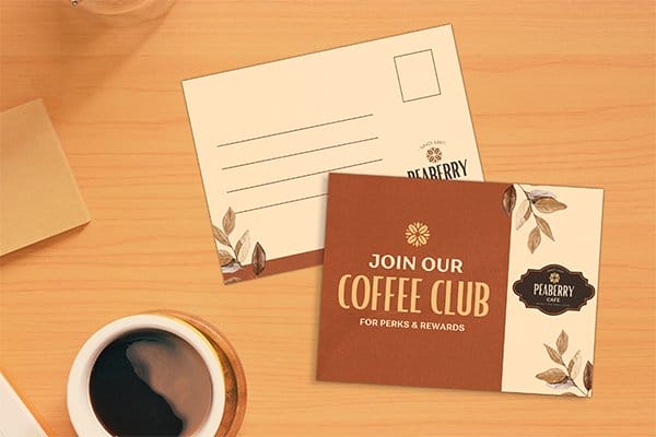 The front and backside of a postcard is shown on a wooden table; the postcard is branded Peaberry Café and reads, 'JOIN OUR COFFEE CLUB.'