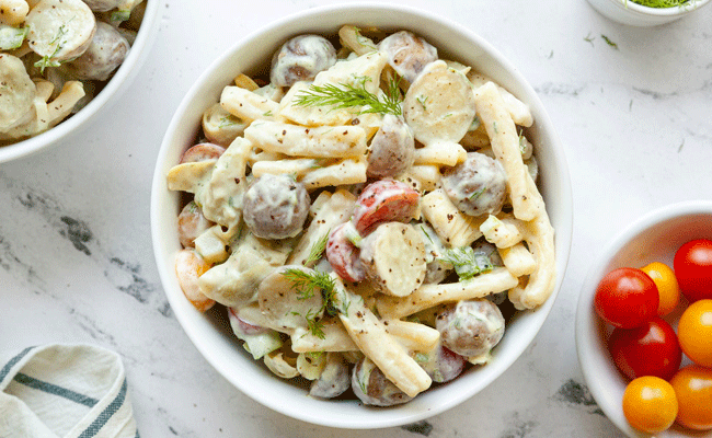 Potato and Artichoke Heart Pasta Salad in a white bowl on a marble table top next to a small white bowl of cherry tomatoes