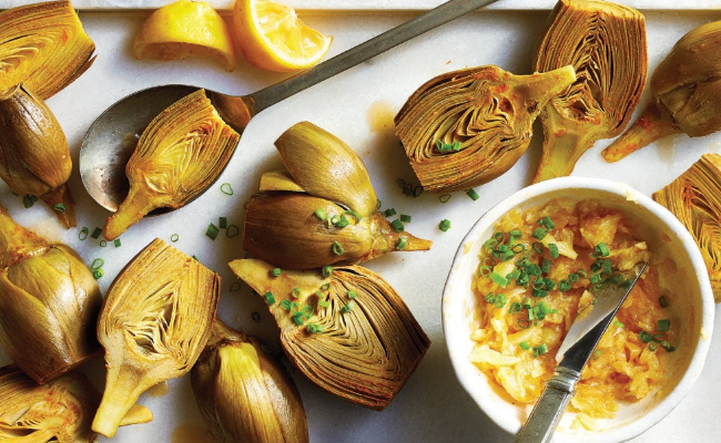 Lemon-Garlic Roasted Artichokes on a white platter with lemon wedges and a small white ramekin of crushed garlic