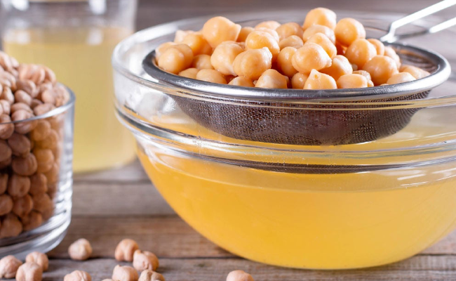 A mesh strainer full of chickpeas is placed over a glass bowl where aquafaba has drained down into it. A glass of dried chickpeas sits to the lefthand side.