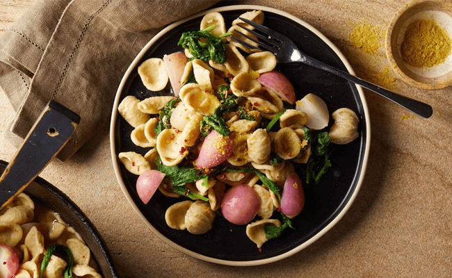 Root to Leaf Radish Pasta in a black bowl on a wooden table with a brown cloth napkin