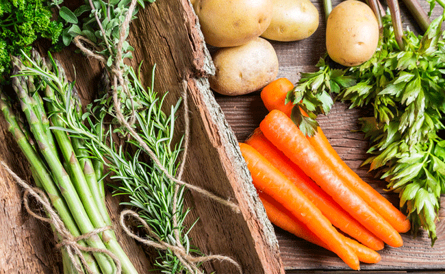 A bunch of fresh asparagus and and another of rosemary tied with string in a rustic wooden box on a wood table next to six carrots, four potatoes and rhubarb