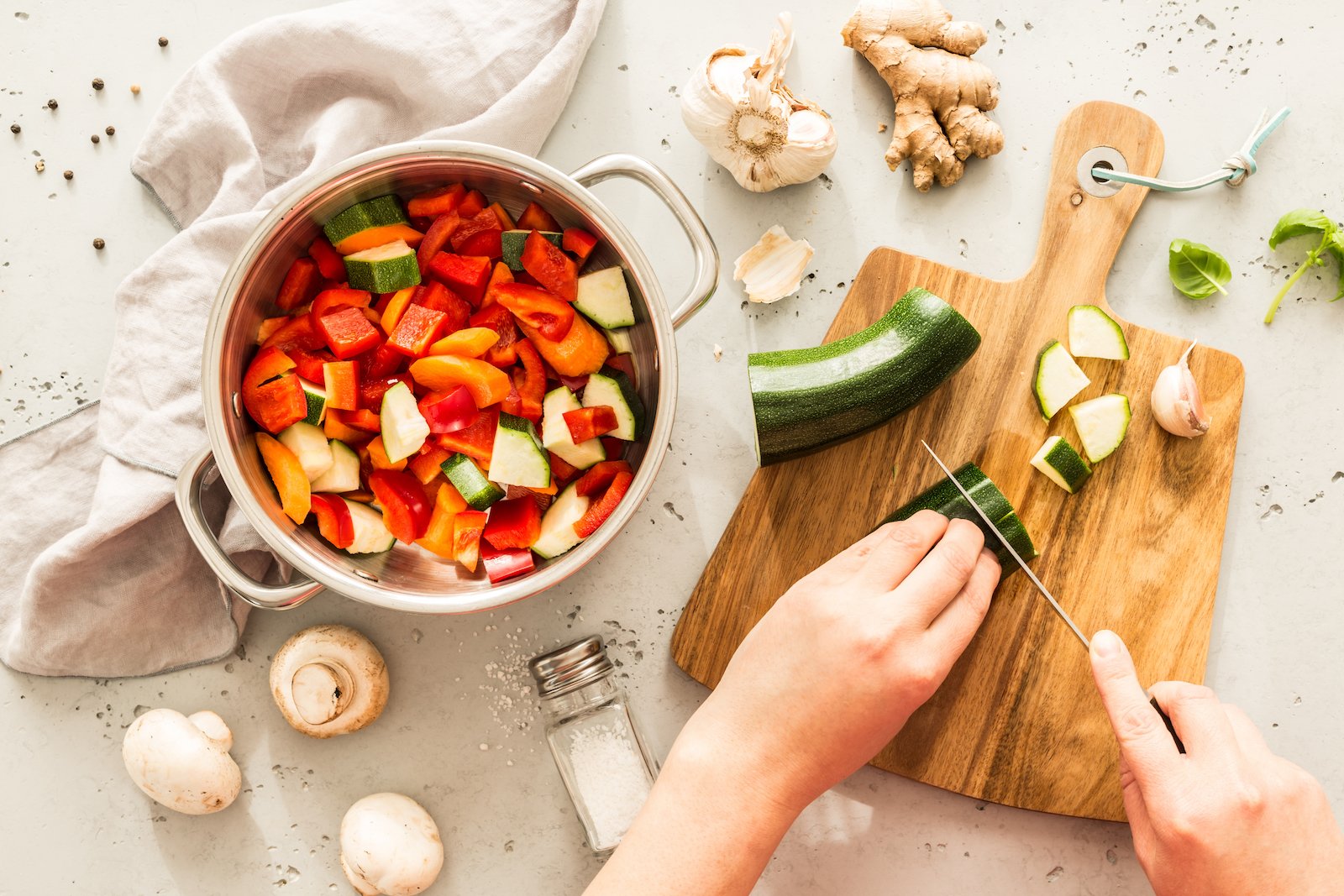 Birds Eye view of a pair of hands chopping a zucchini on a wooden chopping board next to a large pot filled with colorful veggies