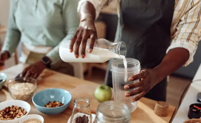 In a kitchen with a wooden countertop, a man and woman arrange bowls of healthy plant based ingredients, with the man pouring vegan milk into a smoothie blender