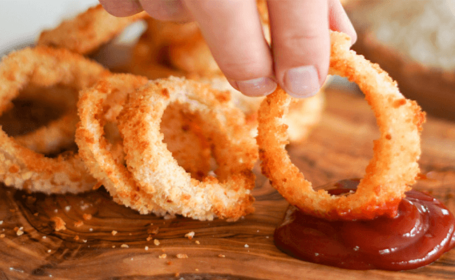 A hand dips an oil-free fried onion ring in ketchup