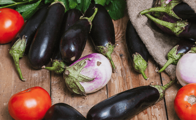 Tomatoes and different varieties of eggplant spill out of a brown sack on to wooden floorboards